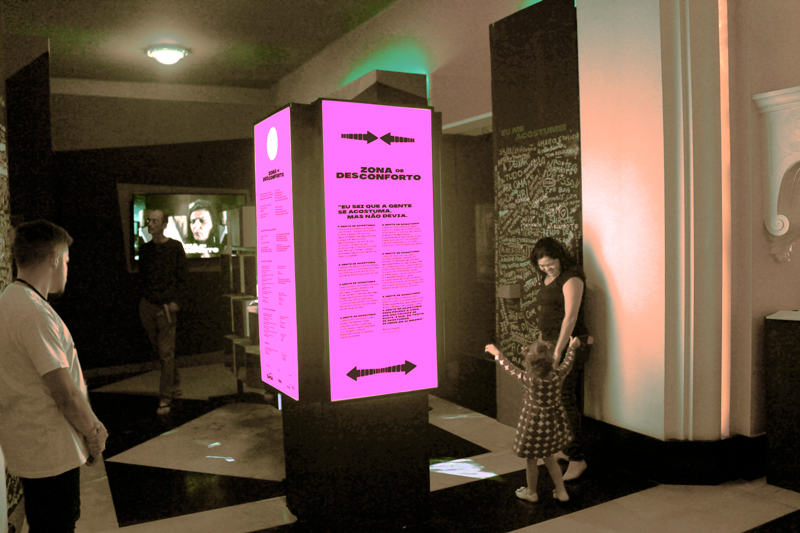 Vista frontal da videoinstalação Zona de Desconforto com foco no totem rosa iluminado e paredes escritas.
